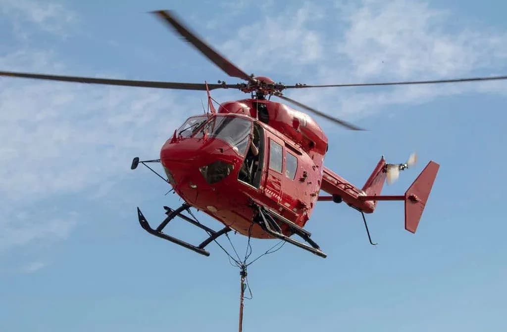 A red helicopter with its main rotor spinning, seen from below, against a blue sky.