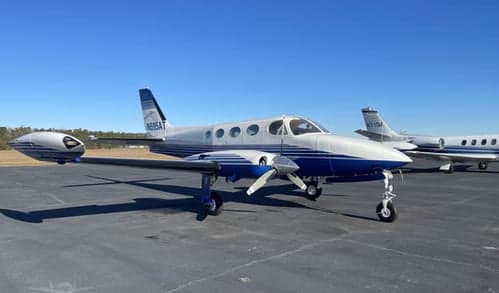 A silver and blue twin-engine propeller aircraft, registration N695AT, is parked on an asphalt tarmac under a clear blue sky, with other aircraft in the background.
