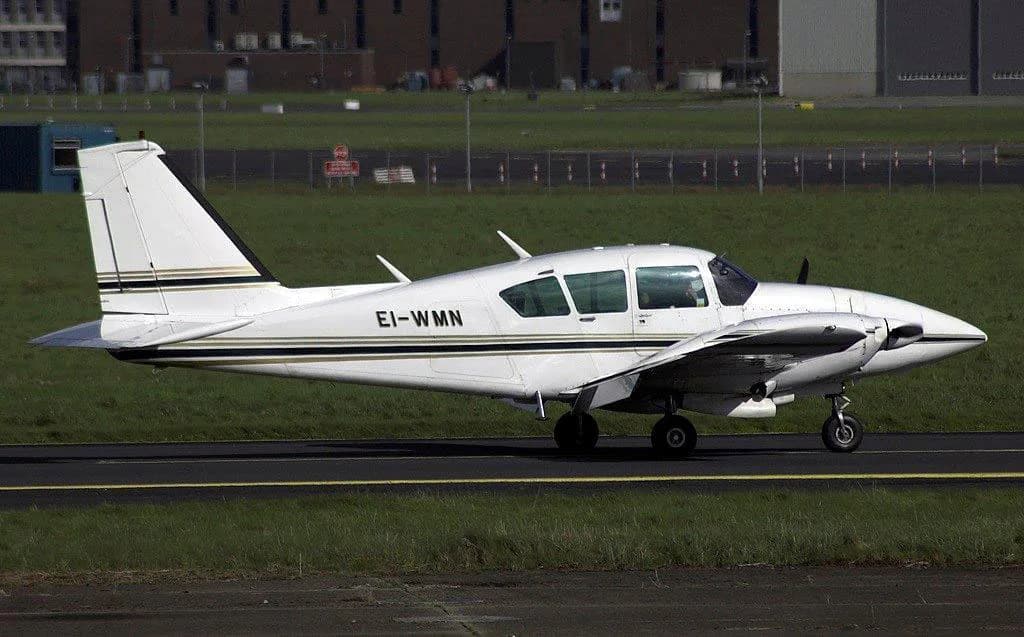 A white twin-engine propeller aircraft with gold and black stripes on the fuselage, registration EI-WMN, on a tarmac.