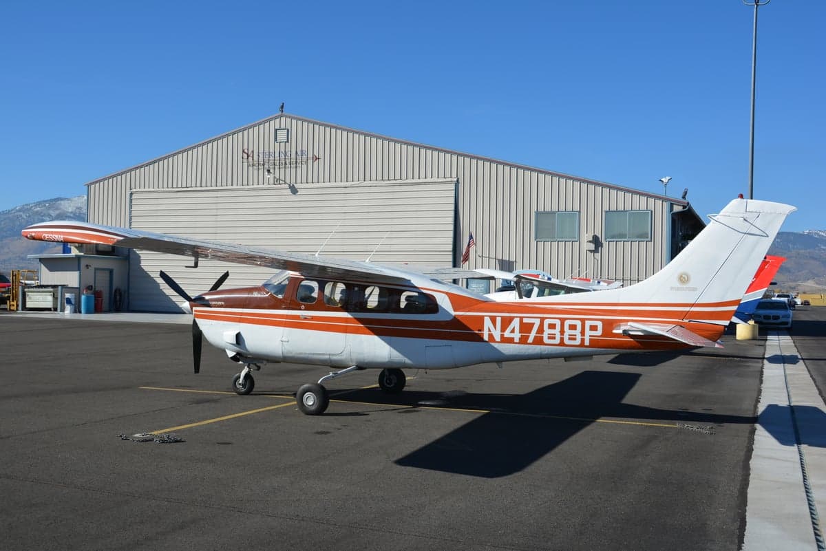 A brown and orange striped Cessna single-engine aircraft, N4788P, parked on an airfield tarmac in front of a hangar with mountains in the background.