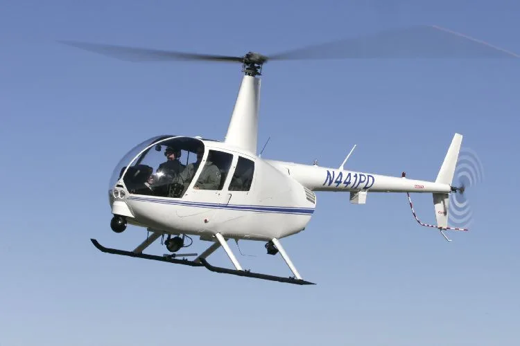 A white Robinson R44 helicopter in flight against a blue sky.