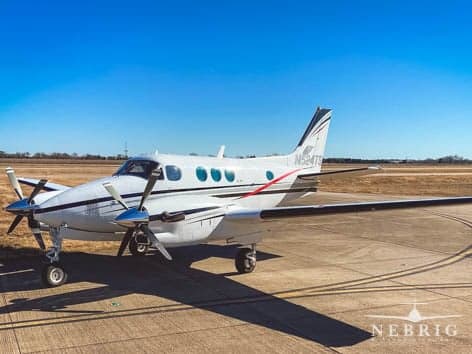 A white twin-engine turboprop aircraft with black and red stripes is parked on a tarmac under a clear blue sky.