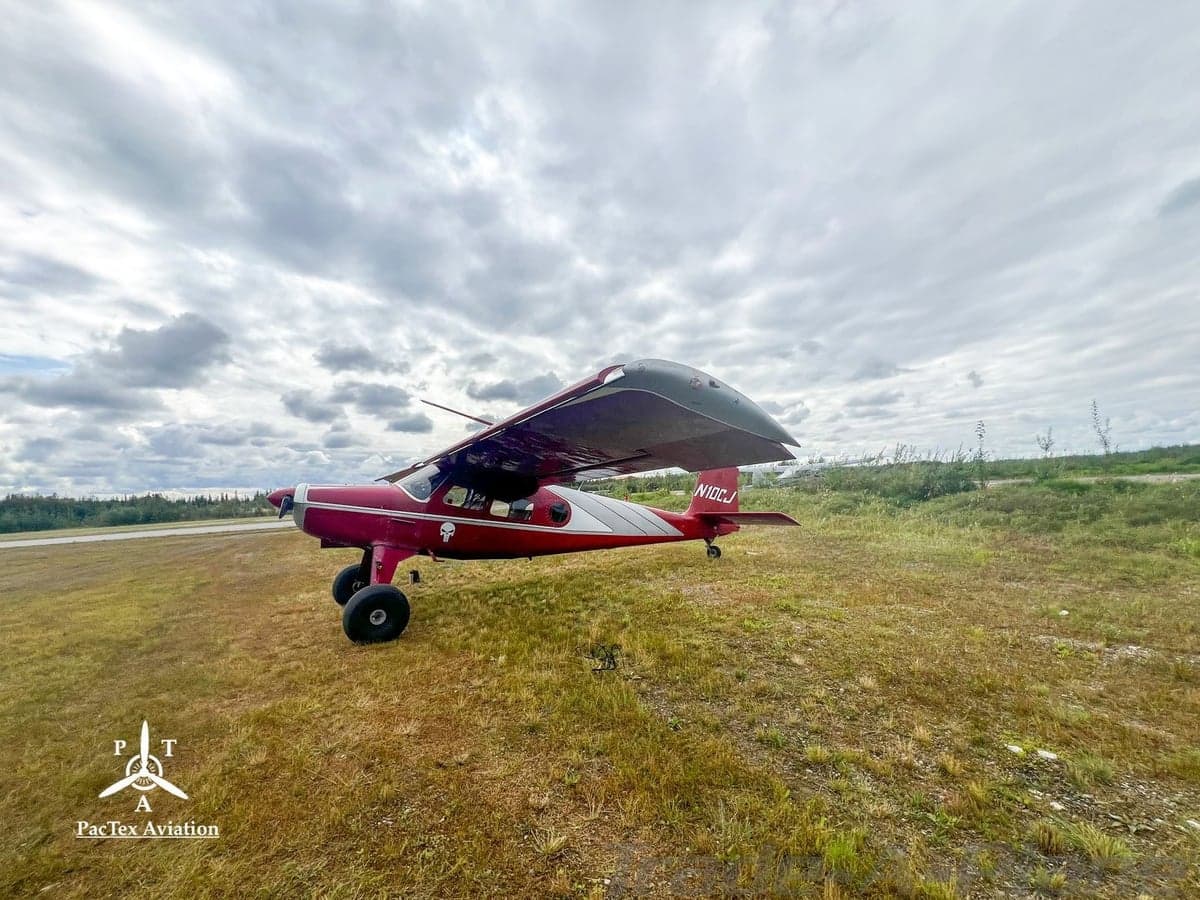 A red and silver single-engine aircraft with large tires, registration N10CJ, parked on a grassy field under a cloudy sky.