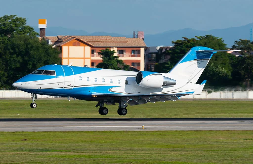 A blue and white Bombardier Challenger 604 business jet is captured mid-air during landing or takeoff, with buildings in the background.