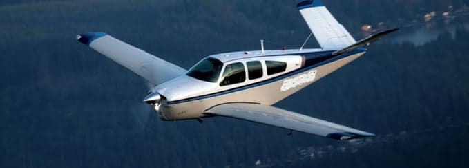 A silver and blue single-engine aircraft with a V-tail flying over a dark, forested landscape.