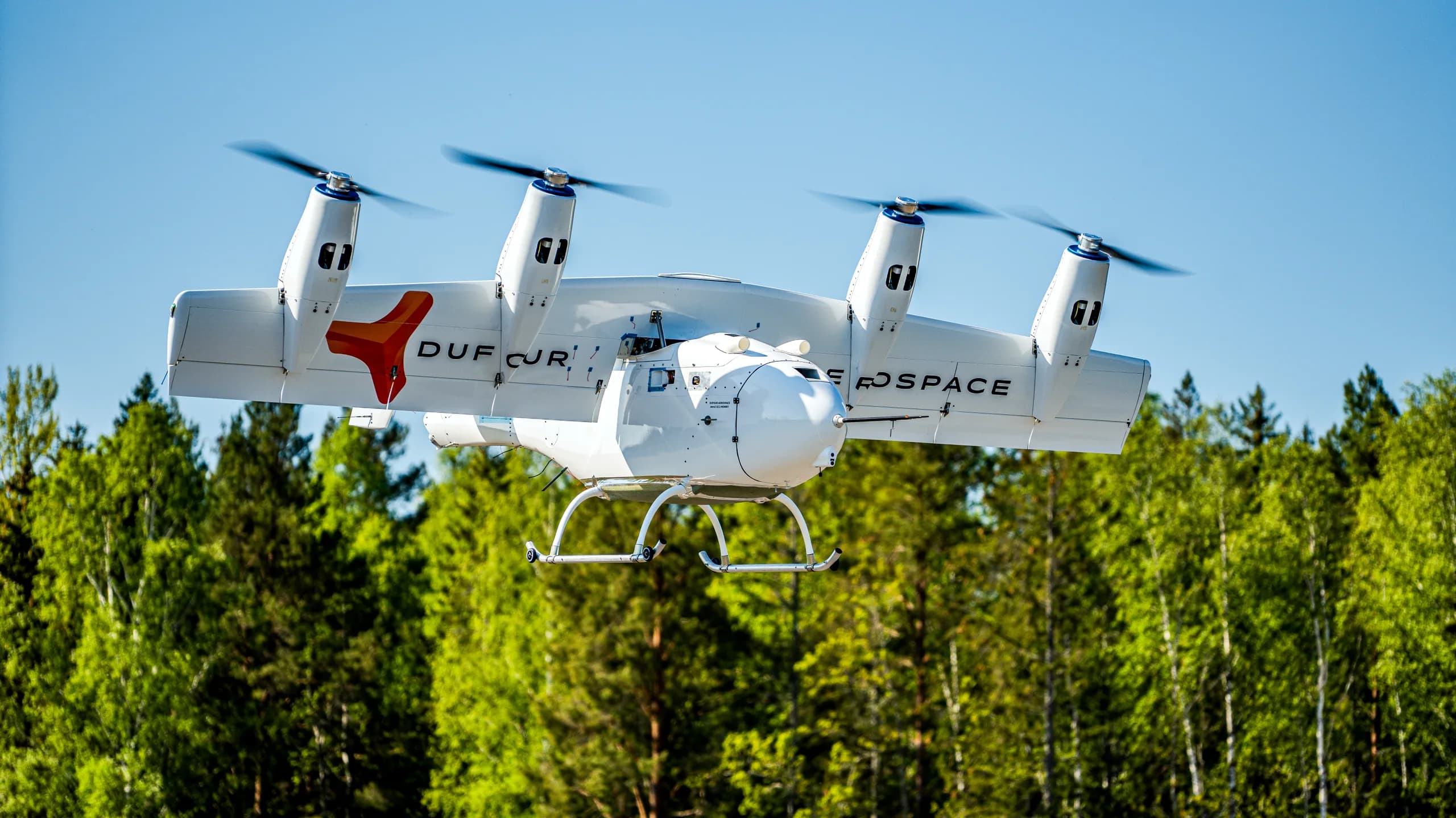 A white Dufour Aerospace eVTOL aircraft with orange accents flying above a lush green forest under a clear blue sky.