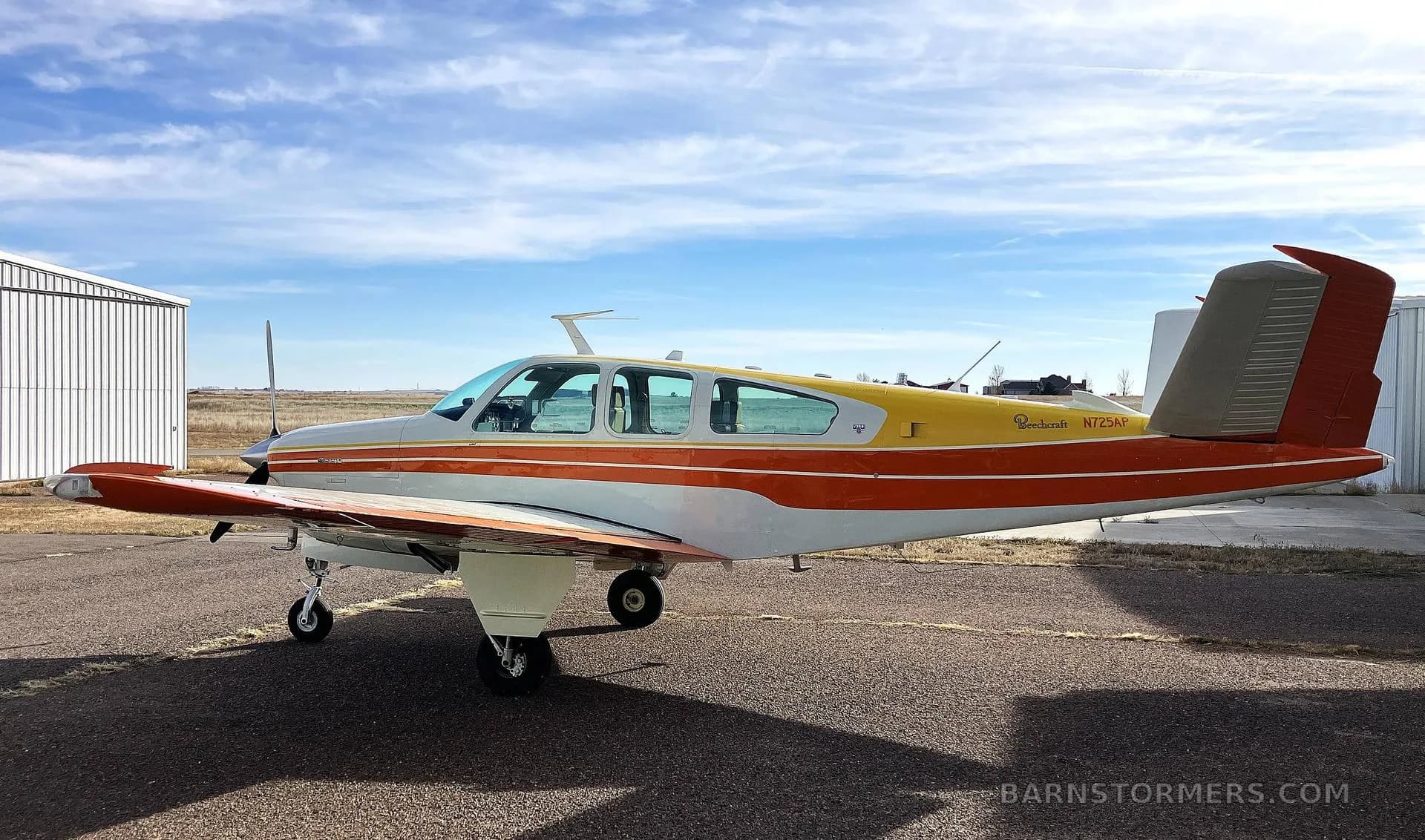 A side view of a white, orange, and yellow Beechcraft Bonanza N725AP parked on an airfield with a hangar in the background.