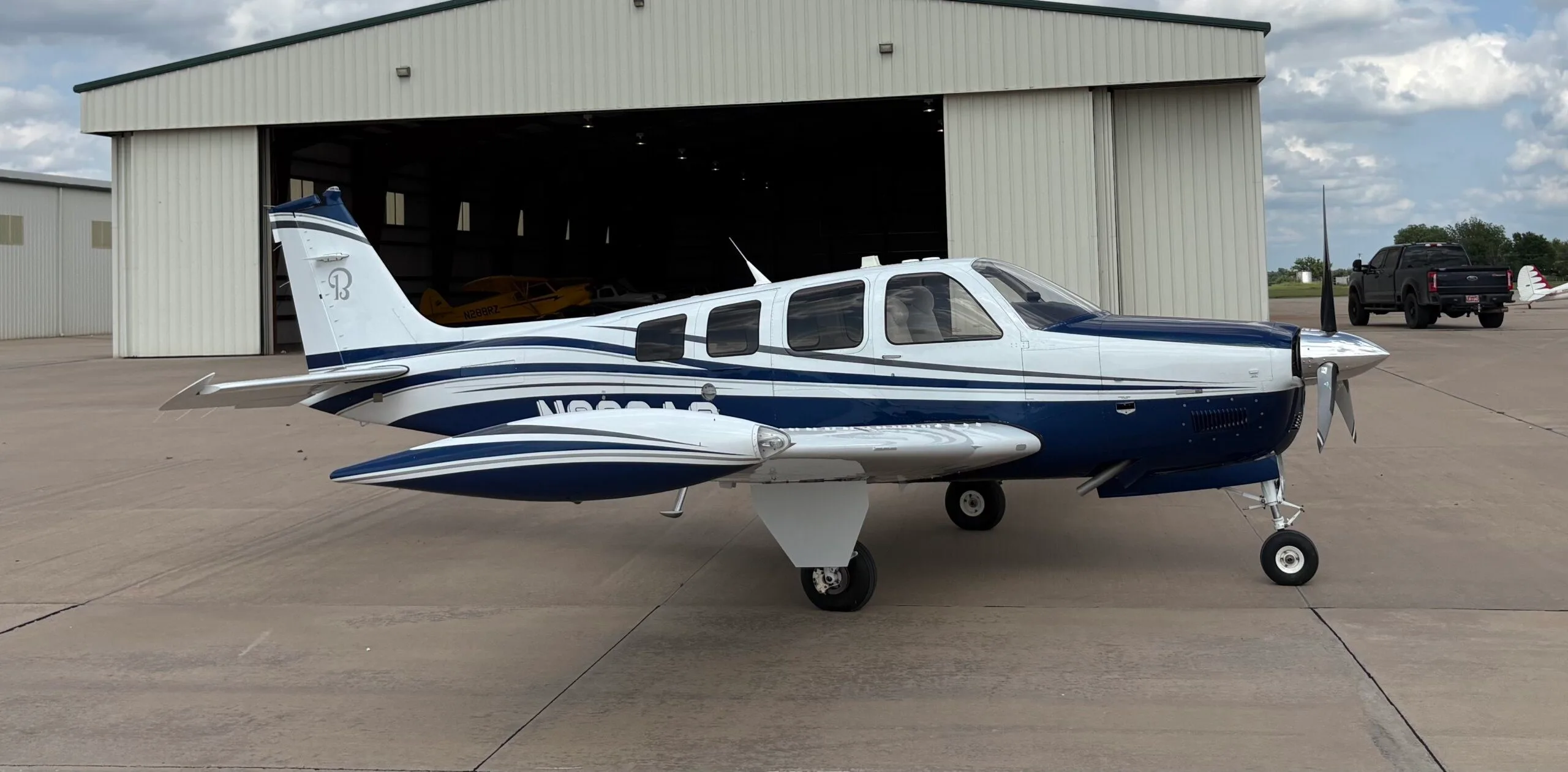 A white and blue Beechcraft Bonanza N686AG is parked on an airfield tarmac in front of an open hangar.