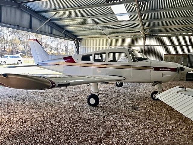 A white and maroon Piper Cherokee 180 parked in a hangar.