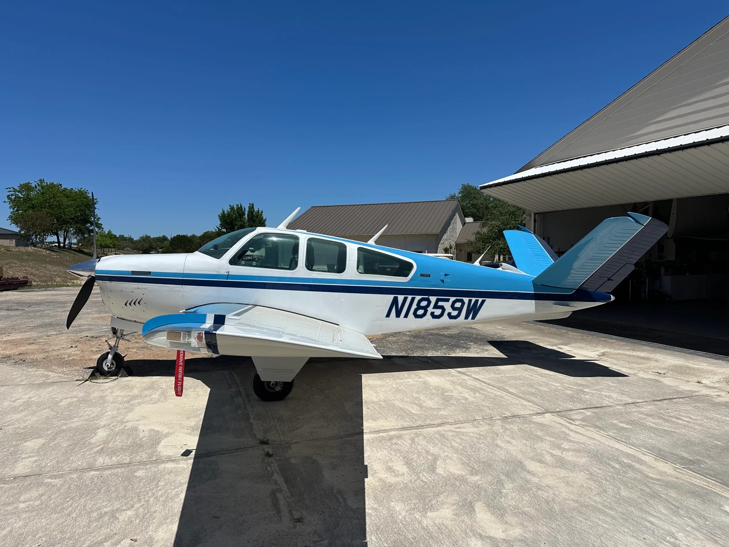 A white and blue Beechcraft Bonanza with registration N1859W is parked on a concrete pad next to an open hangar under a clear blue sky.