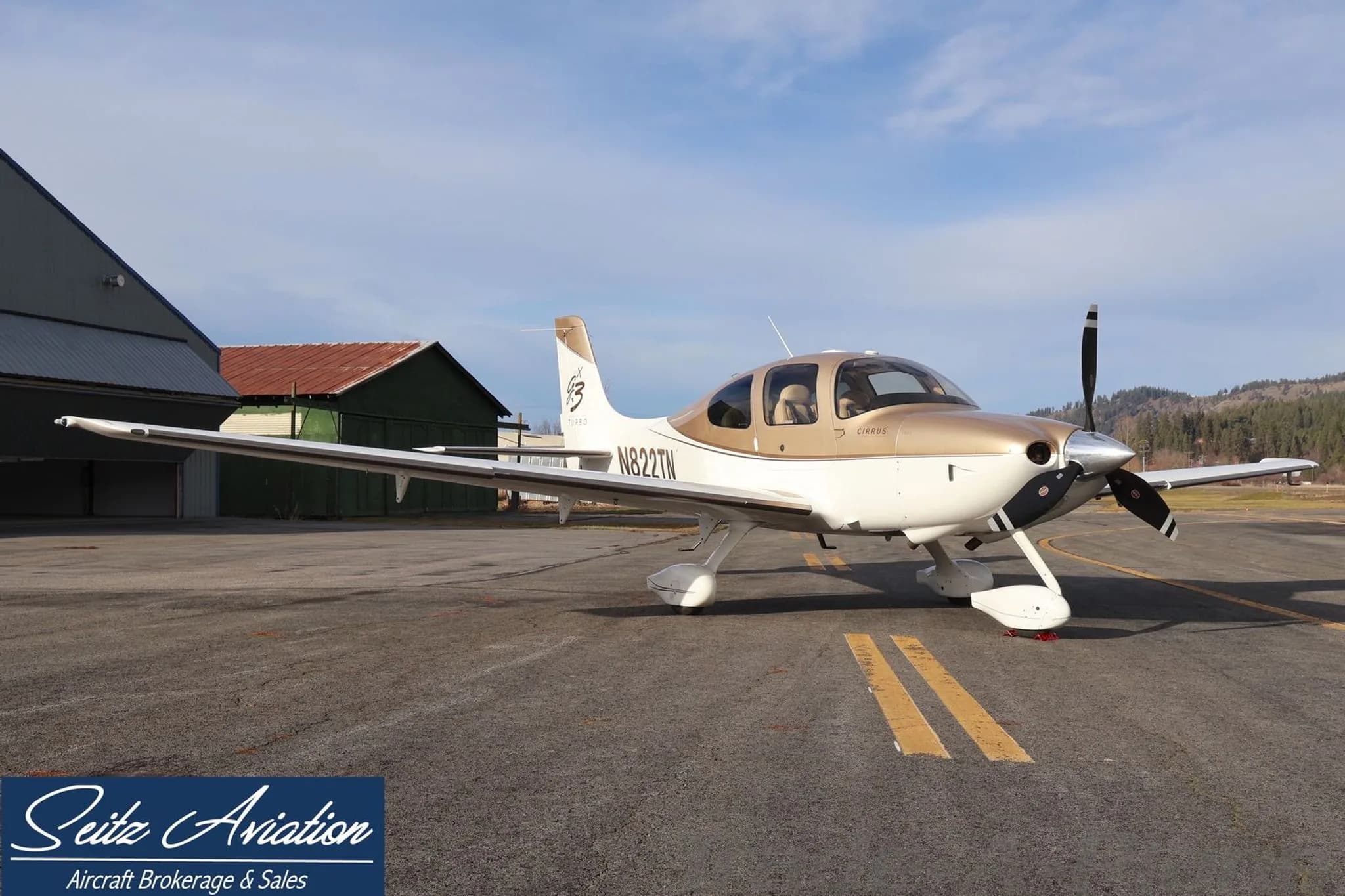 A Cirrus SR22T aircraft with N822TN registration, painted white and gold, parked on an asphalt ramp with hangars in the background.