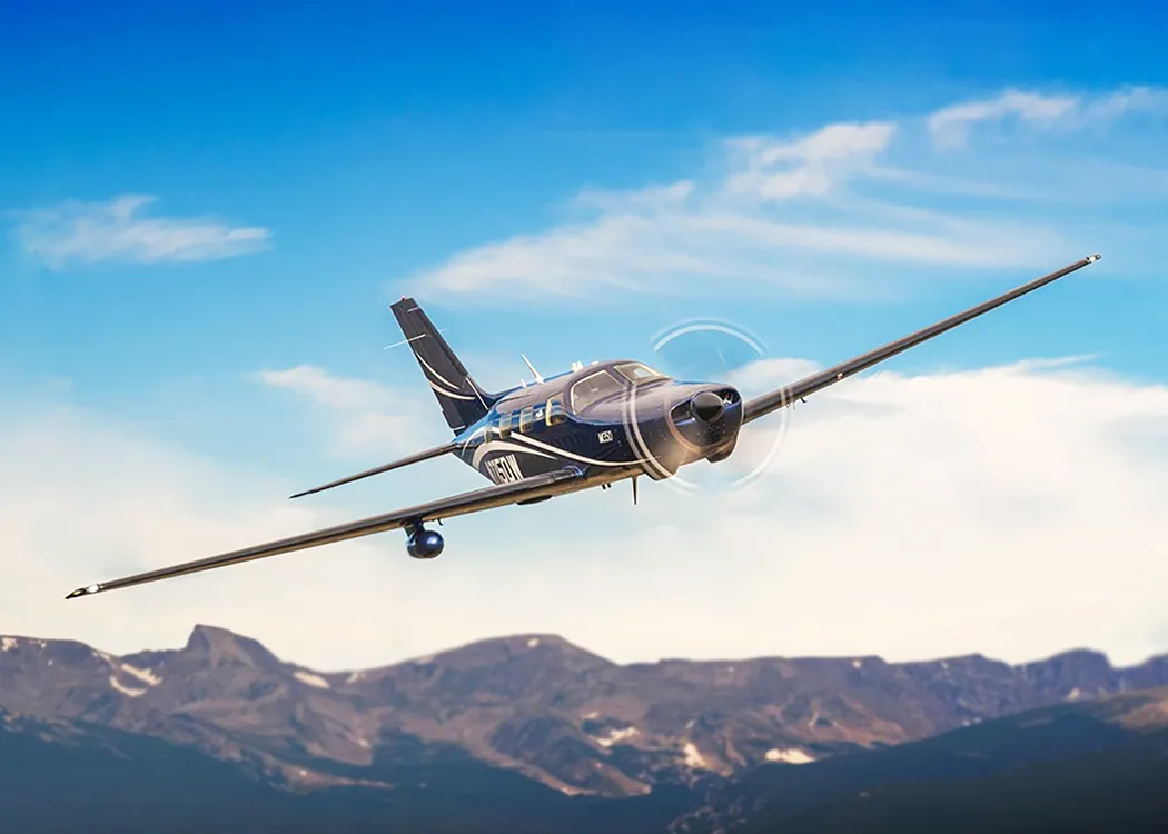 A dark blue Piper M350 aircraft with white and grey accents flying over mountains under a blue sky.