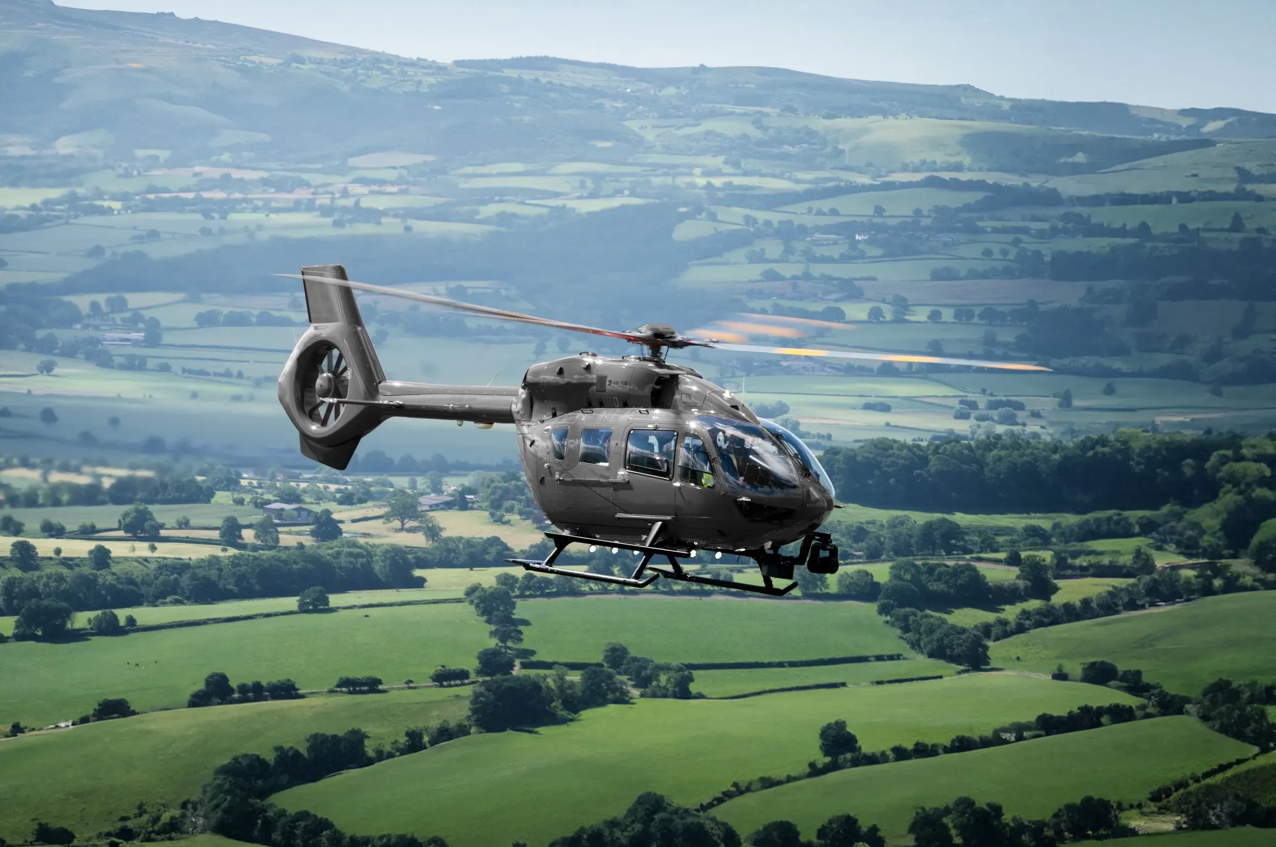 A dark grey Airbus H145 helicopter flies over a lush green, hilly landscape under a bright sky.