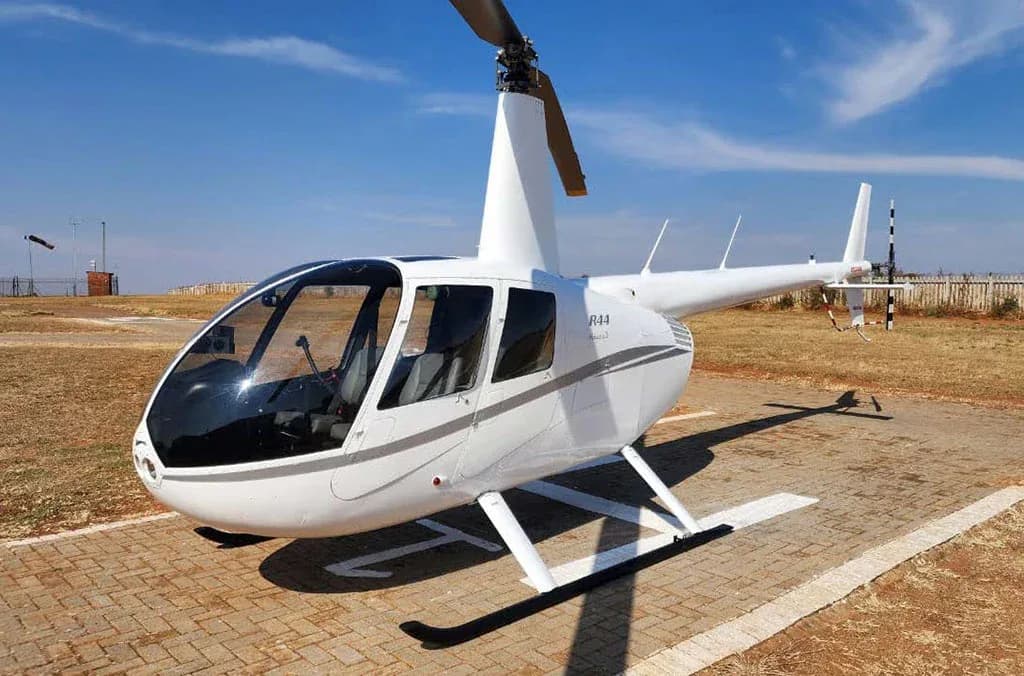 A white Robinson R44 Raven II helicopter with gray stripes sits on a paved helipad under a clear blue sky.