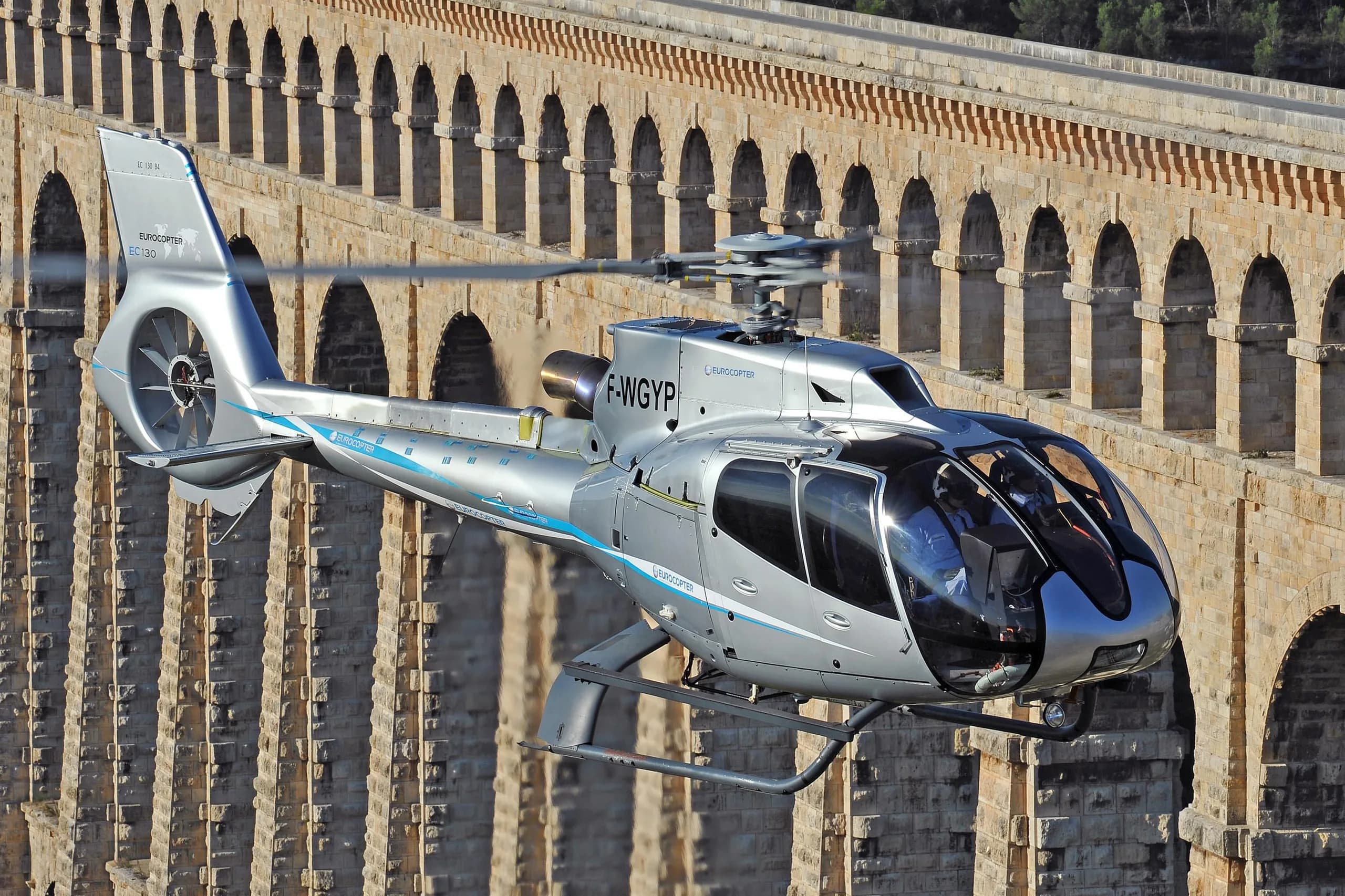 Silver Eurocopter EC135 flying in front of a stone aqueduct.