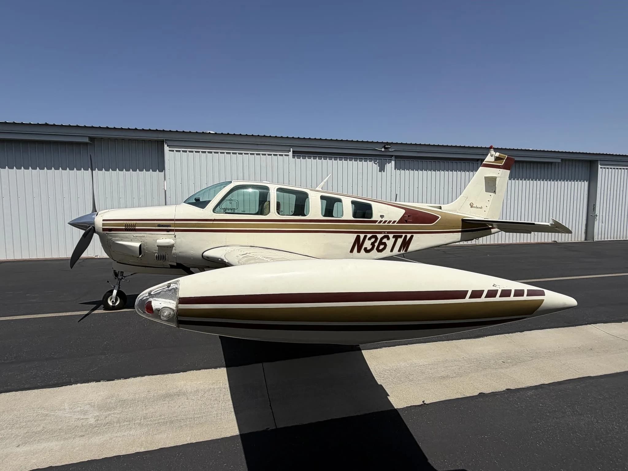 A white and maroon Beechcraft Baron with registration N36TM is parked on an asphalt ramp in front of a hangar under a clear blue sky.
