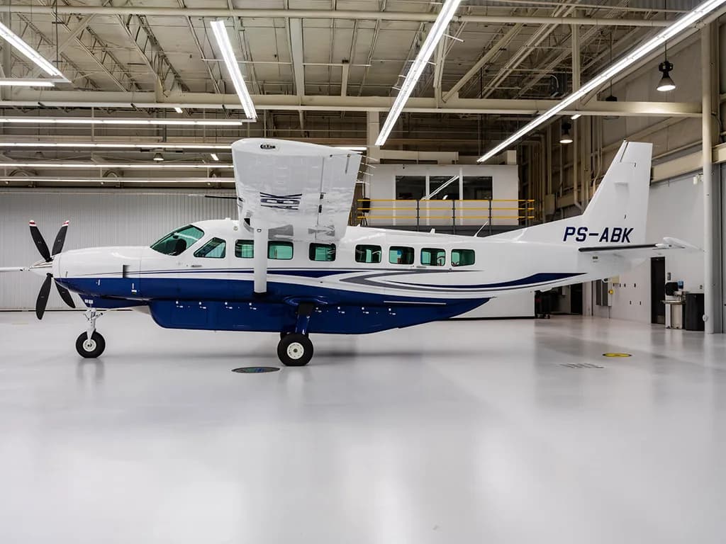 A white and blue Cessna Caravan with registration PS-ABK parked in a brightly lit hangar.