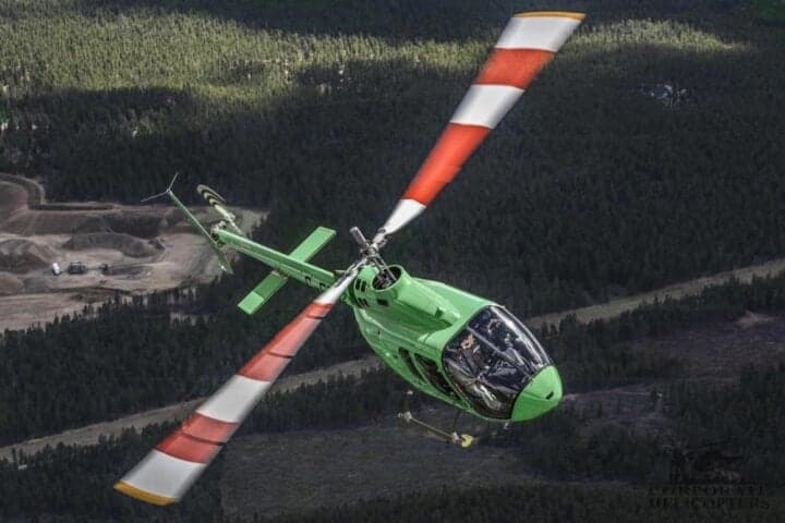 A bright green helicopter with red and white striped main rotor blades flies over a forested landscape with a visible clearing.
