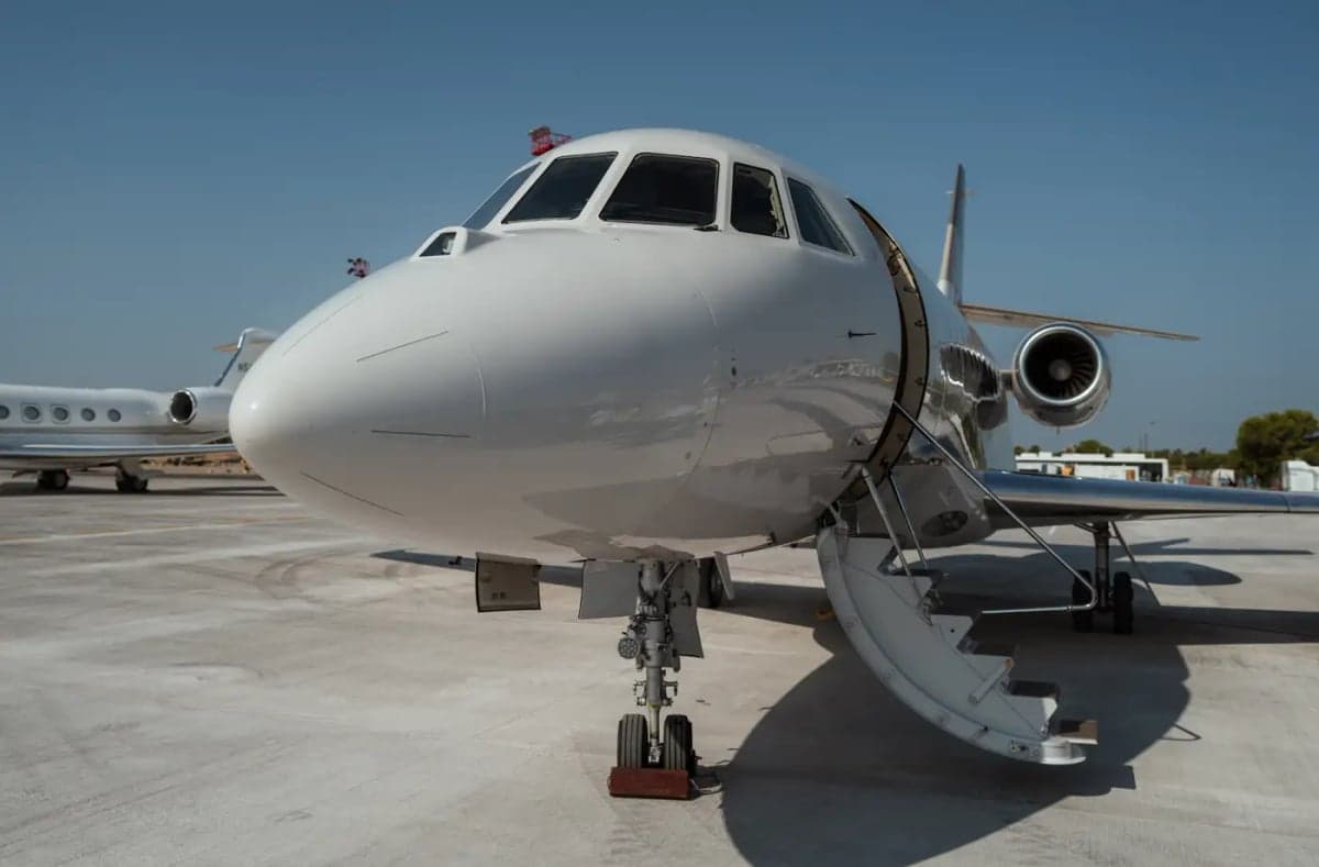 A white Dassault Falcon business jet with its entry door open and stairs extended, parked on an airport tarmac under a clear blue sky.