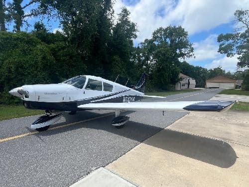 A white and dark blue Piper Cherokee 180 aircraft, registration N7421W, parked on an asphalt surface with trees and a house in the background.