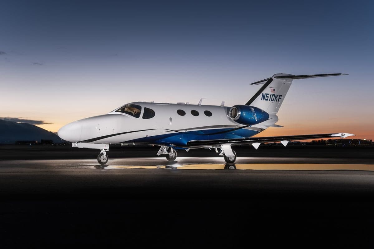 A white and blue Cessna Citation M2 jet (N510KF) is parked on a wet tarmac at dusk, with city lights in the background.