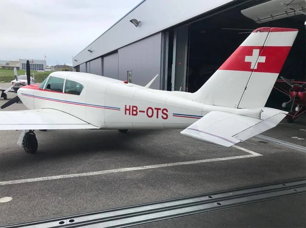 A white single-engine aircraft with red and blue stripes and a Swiss flag on the tail, parked outside a hangar.
