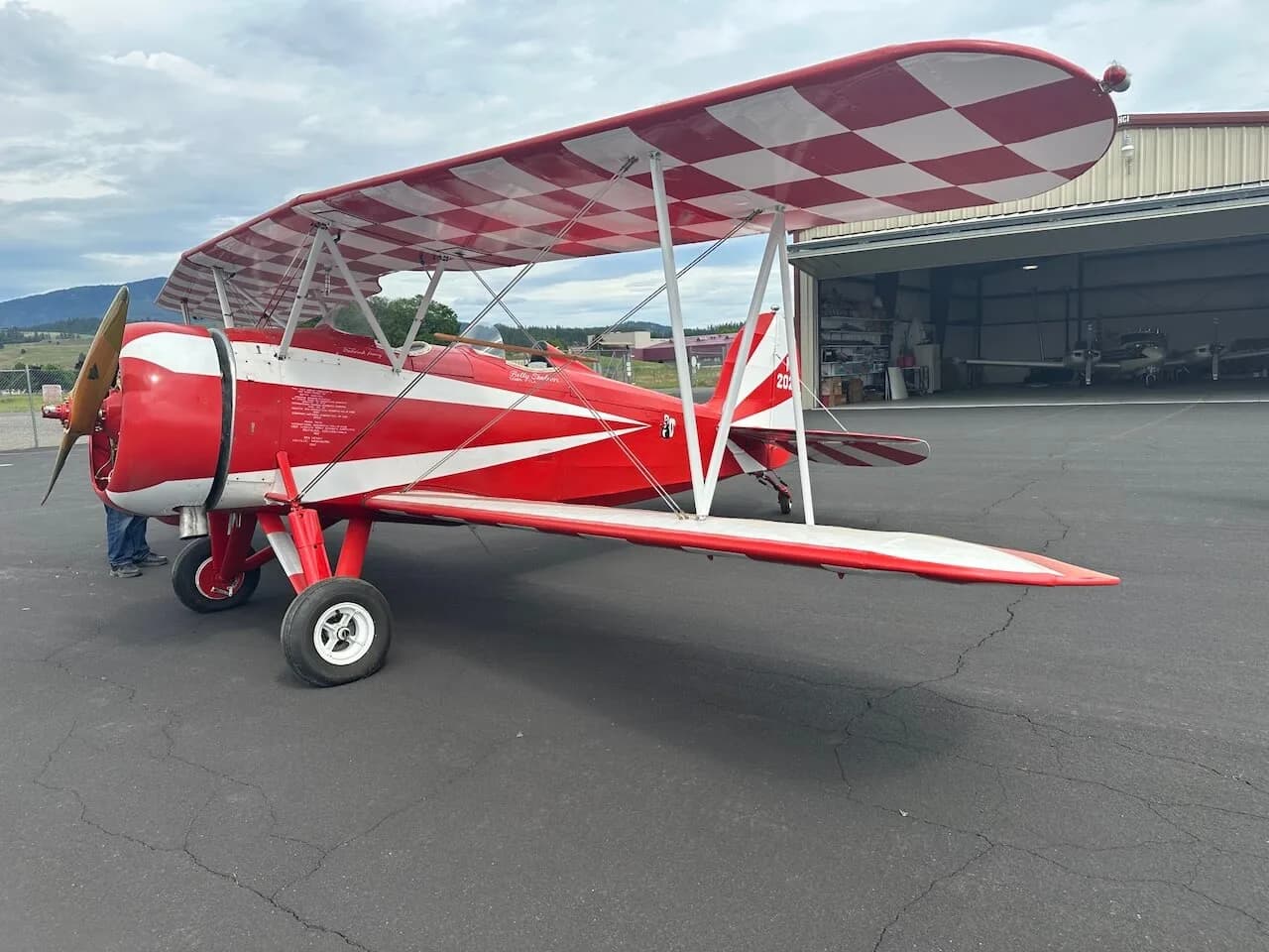 A red and white biplane with a checkered upper wing parked on an asphalt apron in front of a hangar.