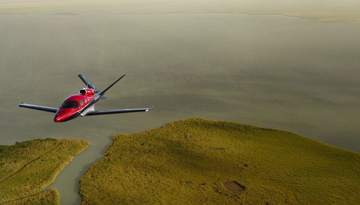 A red and black Cirrus Vision Jet flies over a green landscape.