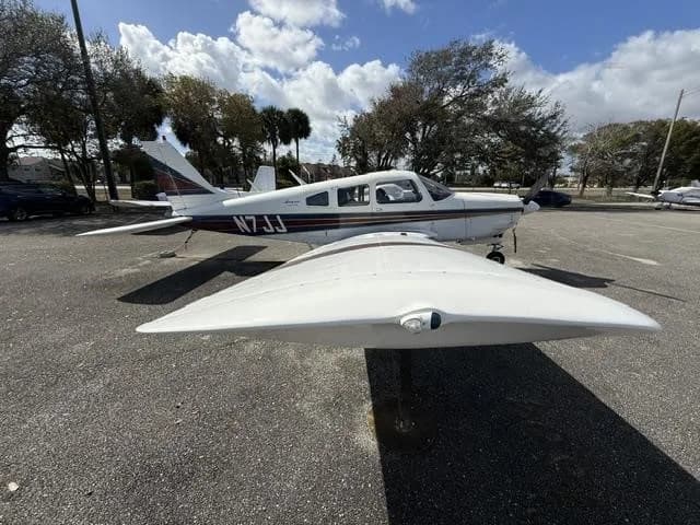 A white and brown Piper Cherokee aircraft, tail number N7JJ, parked on an asphalt lot under a partly cloudy sky.