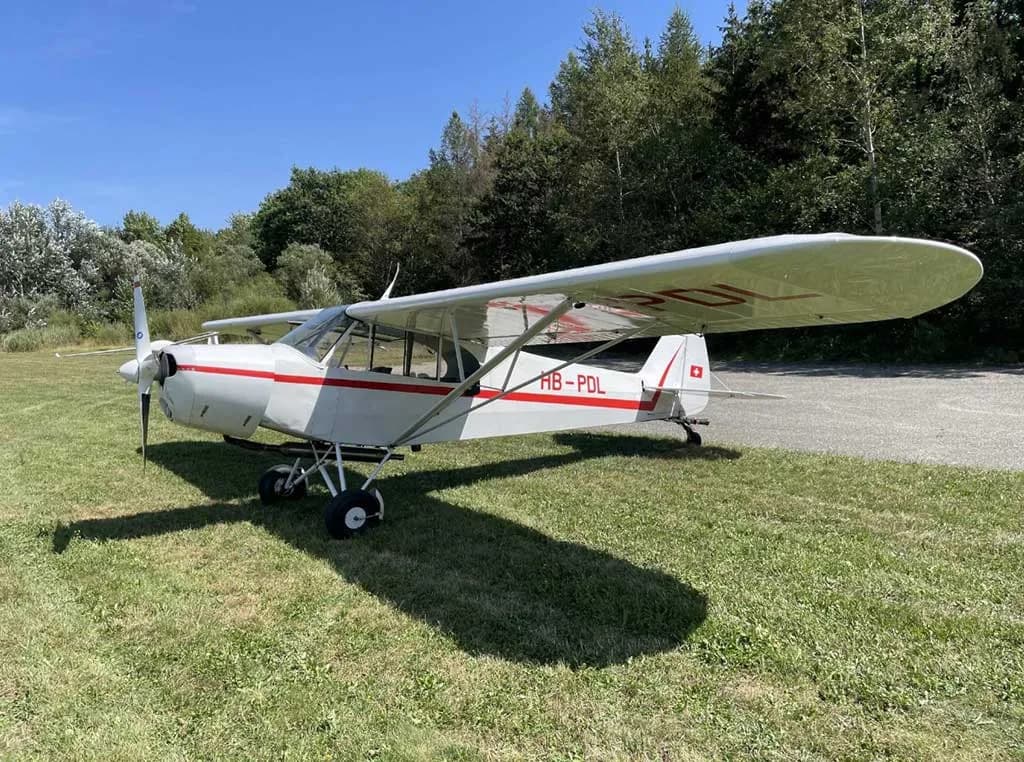 A white single-engine high-wing aircraft with red stripes and Swiss registration HB-PDL parked on a grassy field.