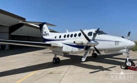 A white and blue Beechcraft King Air twin-turboprop aircraft parked on a tarmac next to a hangar under a clear sky.