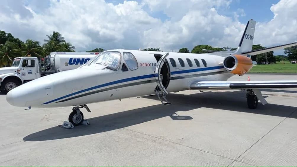 A white business jet with blue and grey stripes, registration CC-AYR, parked on an airfield with its door open and a fuel truck nearby.