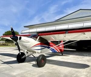 A white and red aircraft is parked in front of a hangar on a sunny day.
