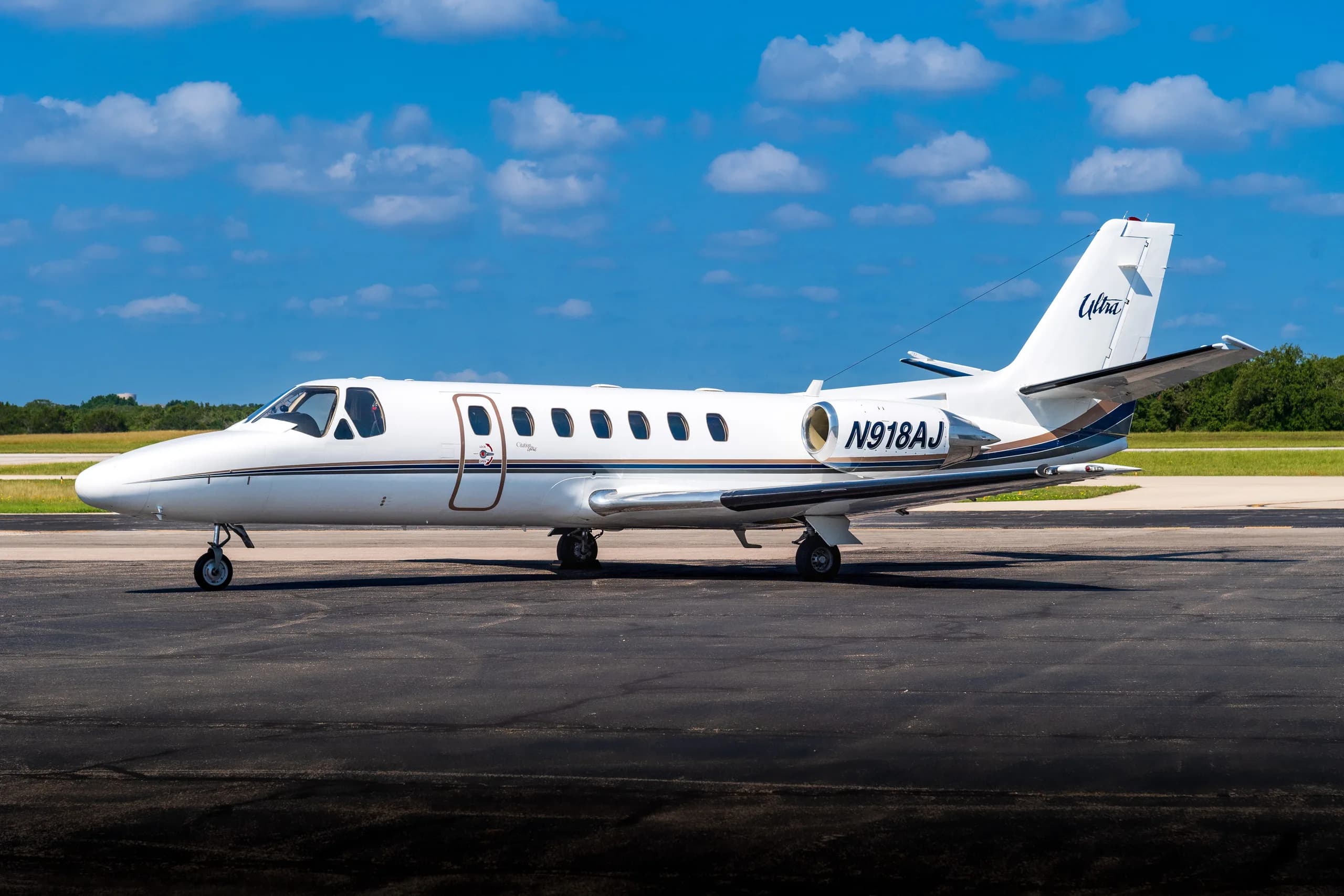 A white Cessna Citation Ultra business jet, registration N918AJ, with blue and brown accent stripes, parked on an airfield under a blue sky.