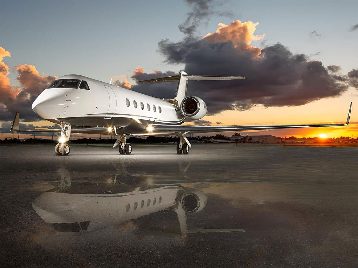 A white Gulfstream G650 business jet with its landing lights on, parked on a wet tarmac at sunset.