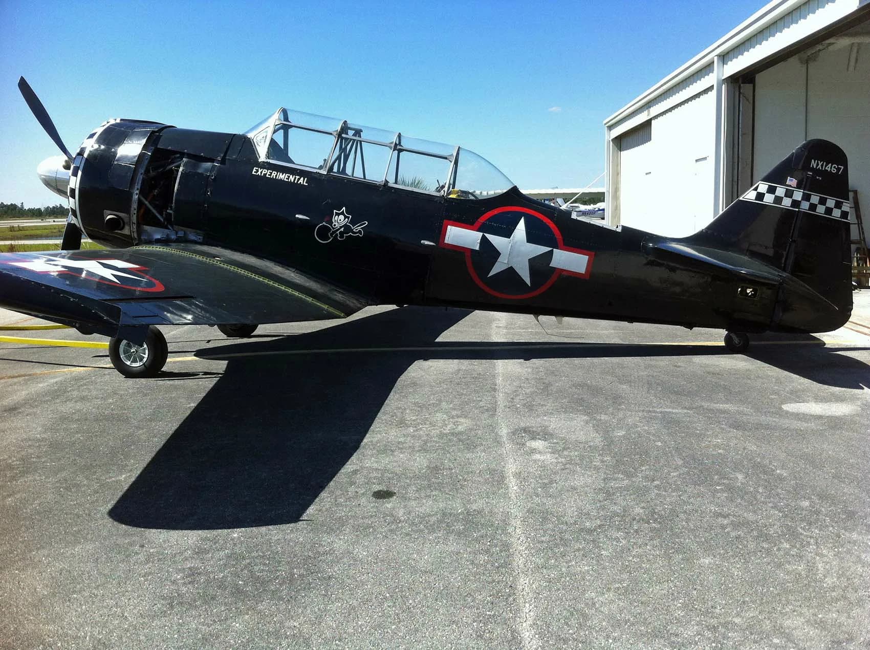 A black warbird aircraft with a checkered nose and US star insignia, registration NX1467, parked on an airfield.