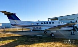 A white twin-engine turboprop aircraft with blue and gold stripes, registration N525SK, parked on a tarmac next to a hangar.