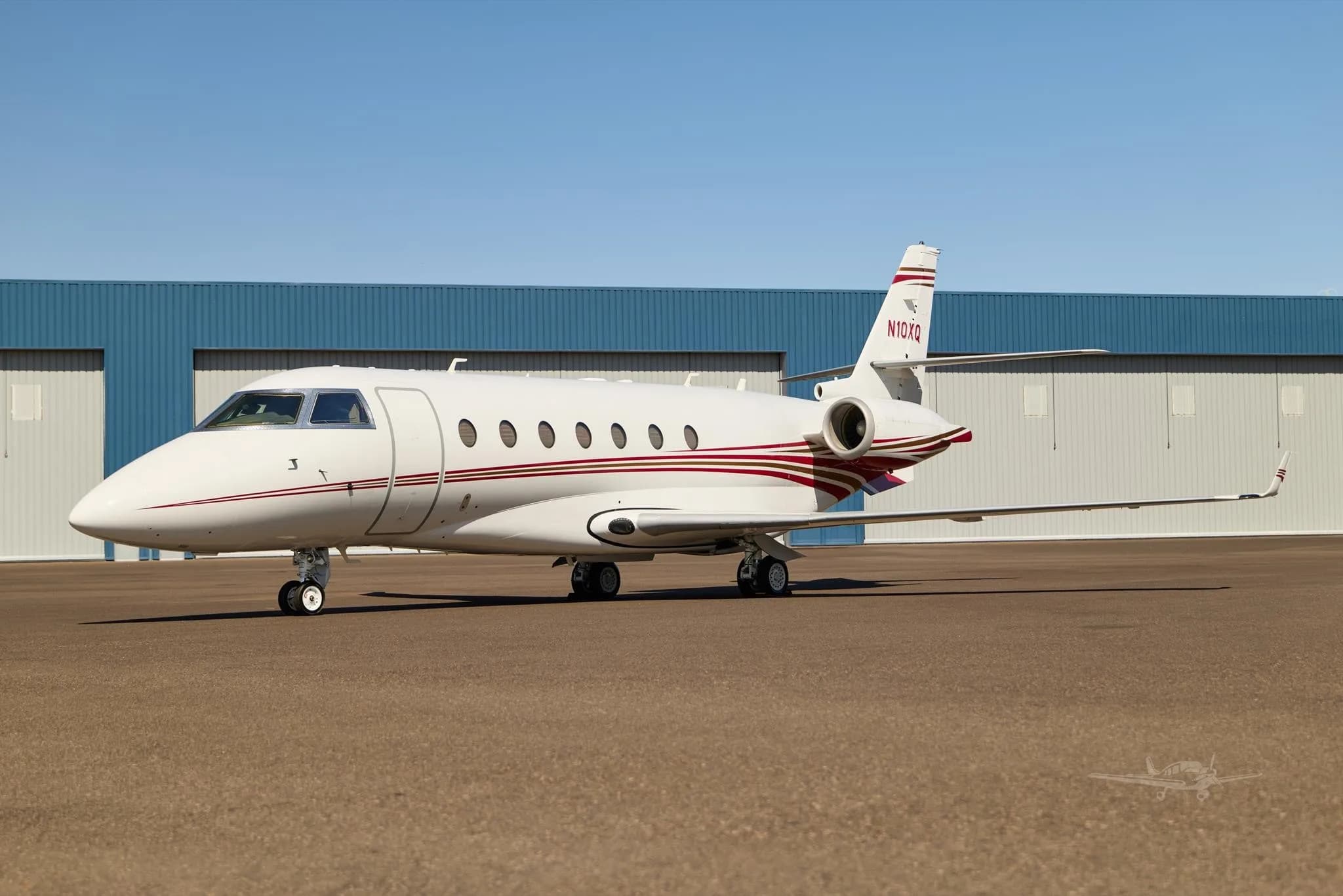 A white business jet with red and gold stripes, tail number N10XQ, parked on an airfield in front of hangars.