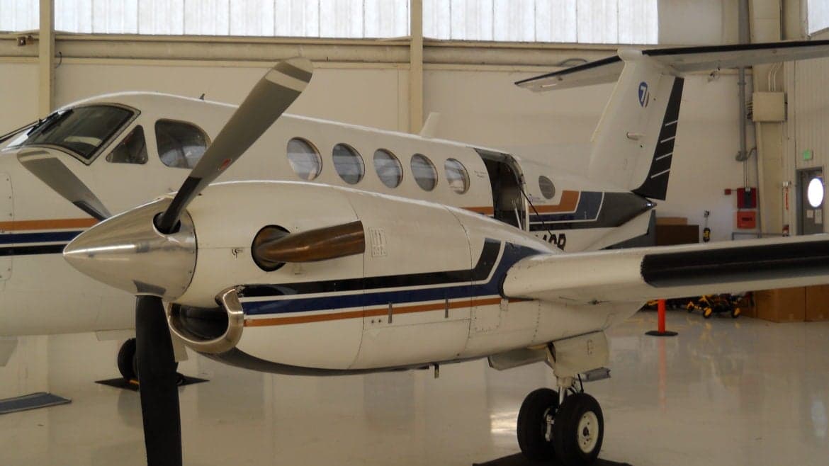 A Beechcraft King Air is parked in a hangar, showing the nose, propeller, and tail section.