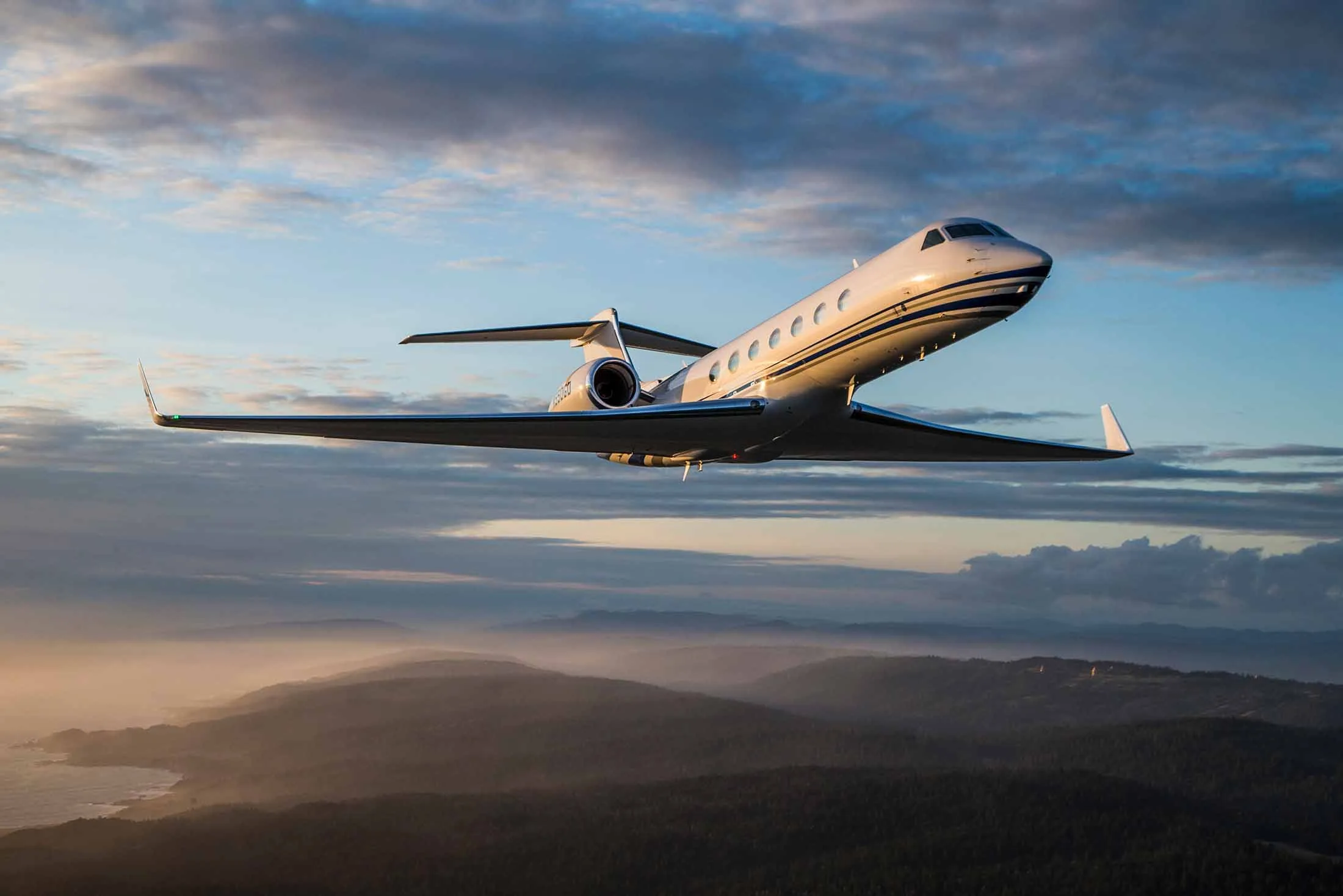 A white Gulfstream business jet with blue stripes flies over a misty, forested coastline at sunset.