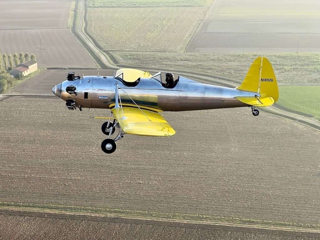 A silver and yellow aircraft, registration N18926, in flight over a rural landscape.