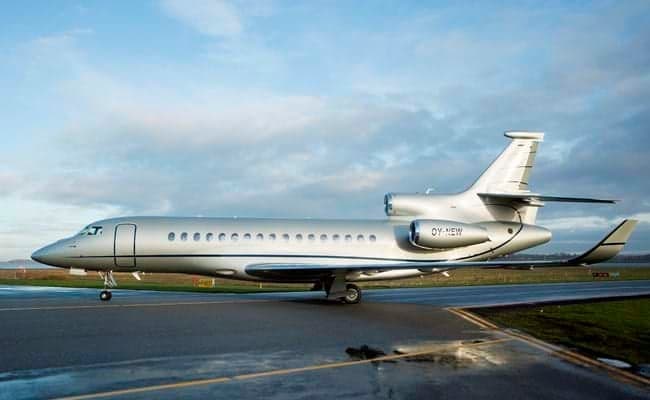 A silver Dassault Falcon business jet with registration OY-NEW is parked on a wet tarmac under a cloudy sky.