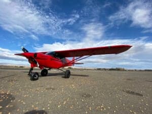 A red Cubcrafters aircraft is parked on a runway under a blue sky with scattered clouds.