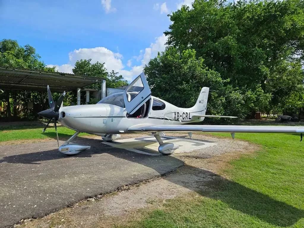 A silver Cirrus SR22 aircraft with its gull-wing door open, parked on a tarmac next to grass and trees under a blue sky.