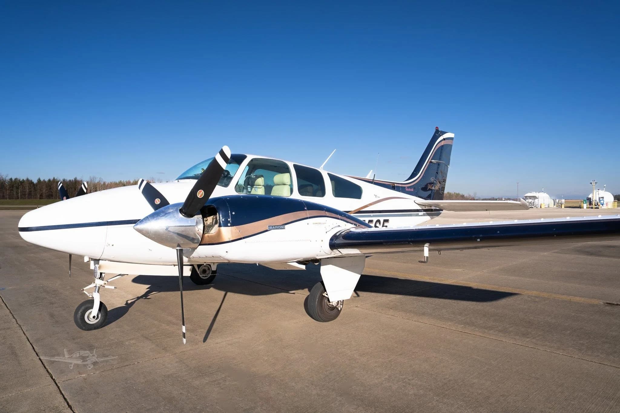 A white and dark blue Beechcraft Baron twin-engine aircraft with tan stripes parked on an airfield under a clear blue sky.