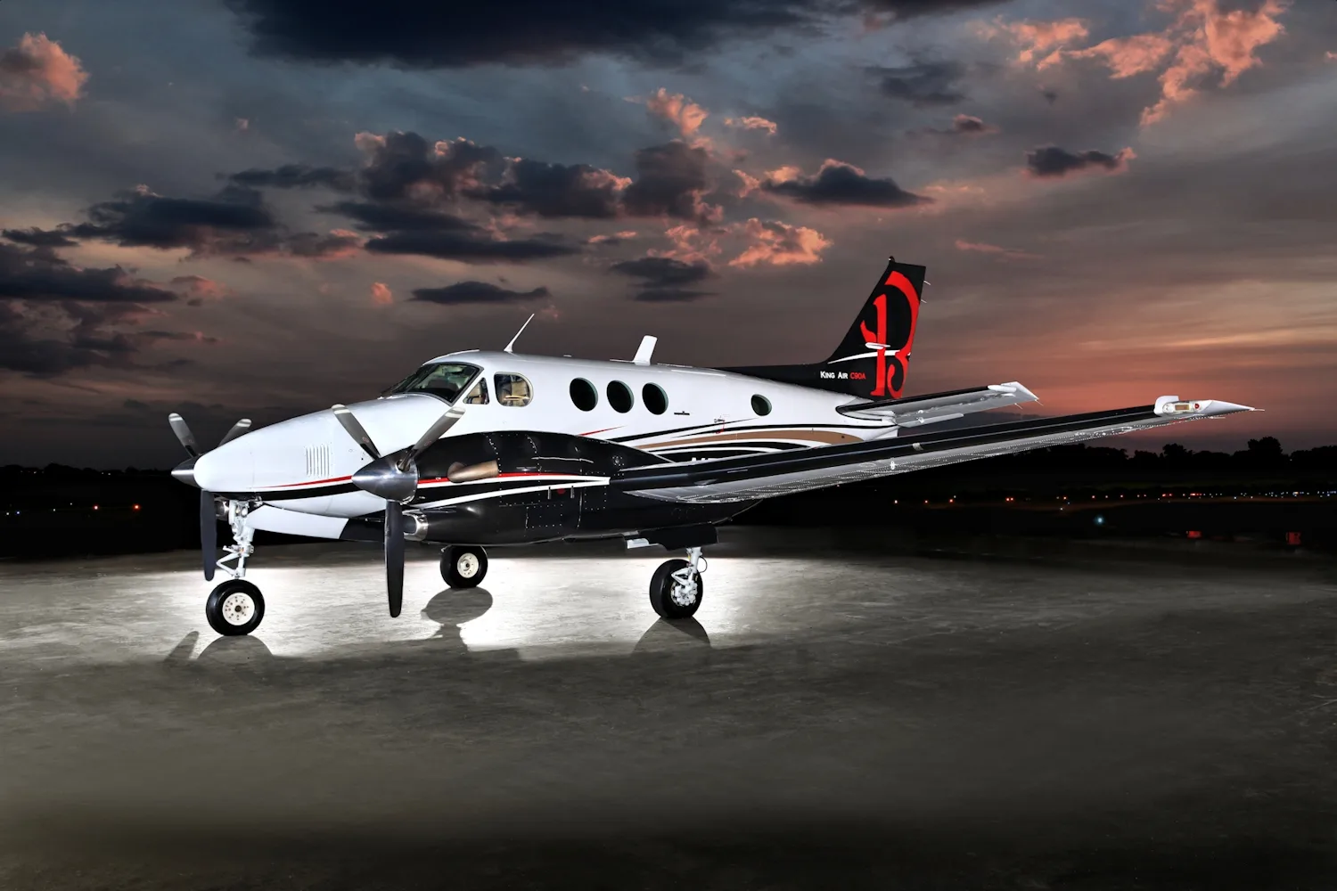 A white and black King Air C90A twin-turboprop with gold accents and a red 'B' tail logo is parked on a tarmac at dusk.