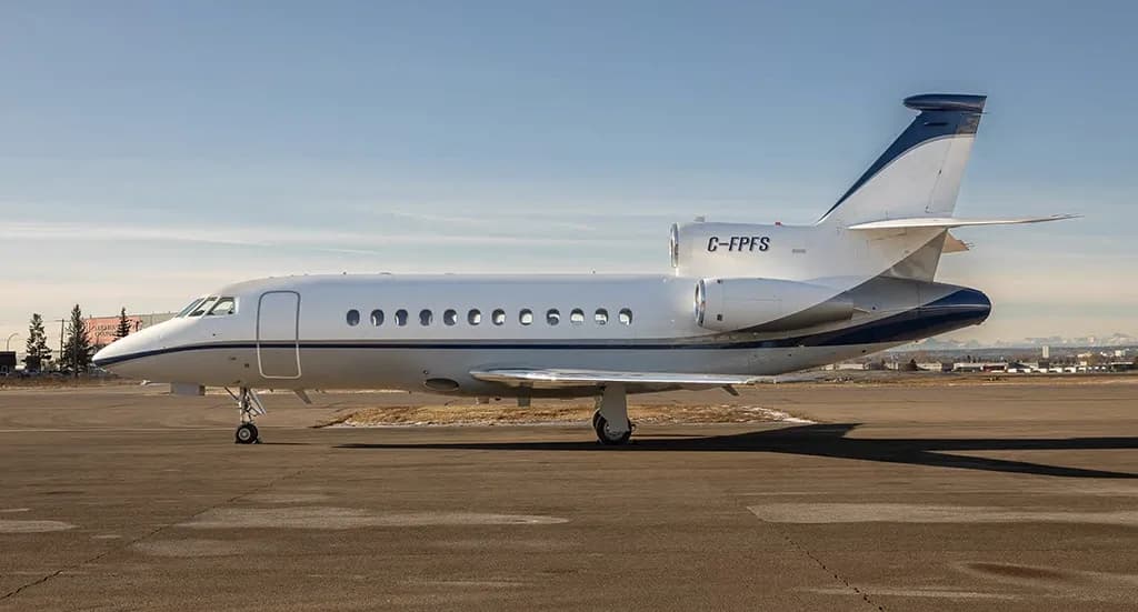 A white and blue Dassault Falcon 900EX business jet with registration C-FPFS parked on an airfield under a clear sky.