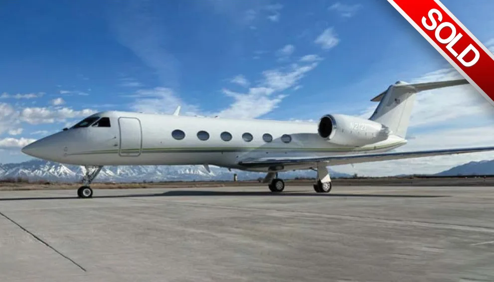 A white Gulfstream business jet with a green stripe, registration N212T, parked on a tarmac with snow-capped mountains in the background.