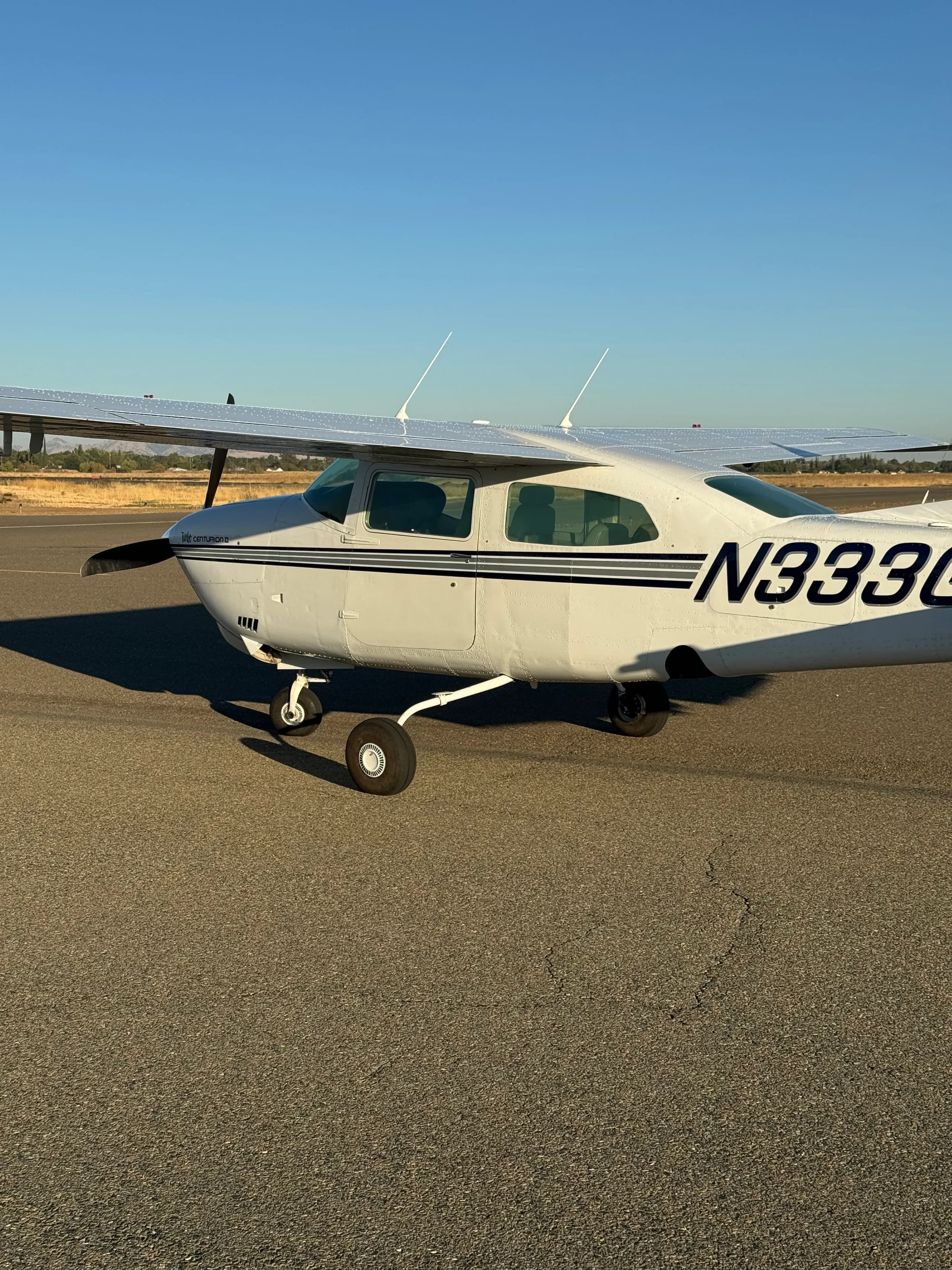 A white Cessna Centurion II with dark blue stripes and registration N333G is parked on an airfield under a clear blue sky.