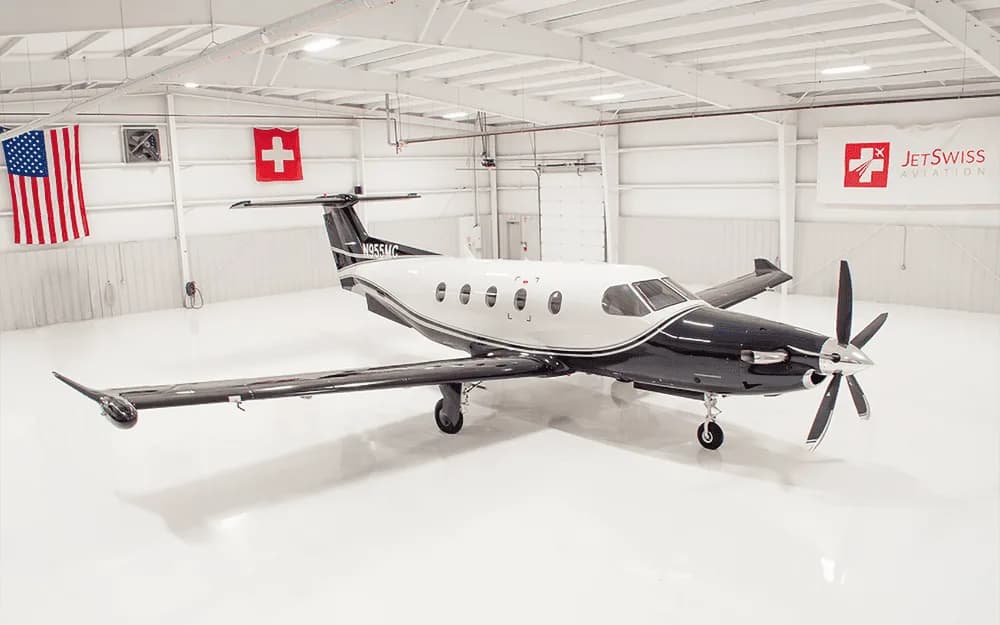 A black and white Pilatus PC-12 turboprop aircraft parked in a brightly lit hangar with US and Swiss flags.